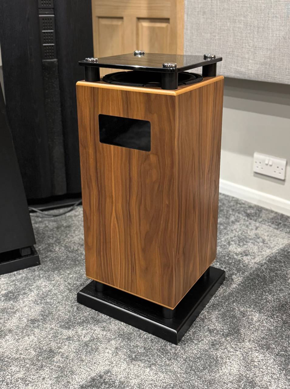 Wood veneer speaker cabinet mounted on a black metal stand with a glass top, placed on a carpeted floor in a room observation