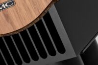 Prophecy-1-014-1080px – NEXT Hi-Fi Close-up of a speaker or audio component with a wooden top panel and black vented grille featuring a small MC logo label toward the edge of the wood panel.