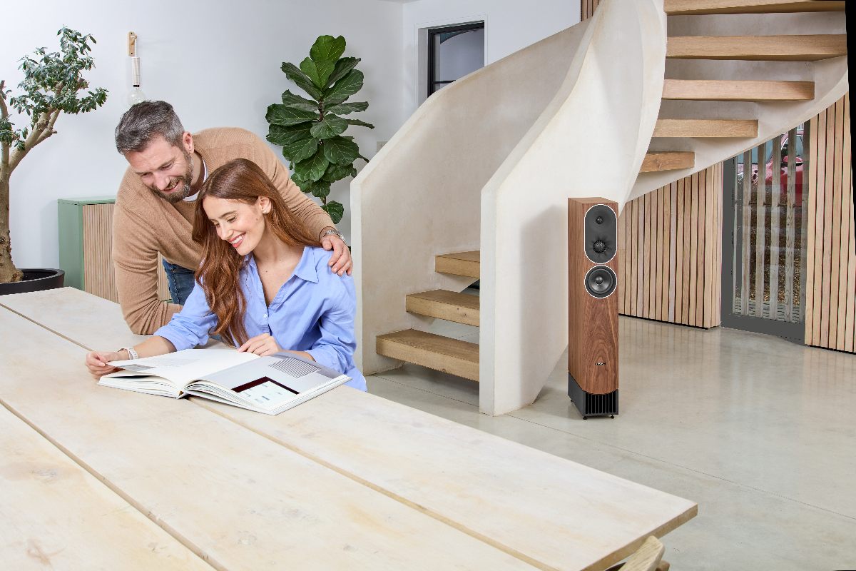 PMC-Prophecy-Loc-1597-1200x800px – NEXT Hi-Fi Два человека рассматривают журнал за столом из светлого дерева. В интерьере их современной квартиры установлены напольные акустические системы PMC prophecy